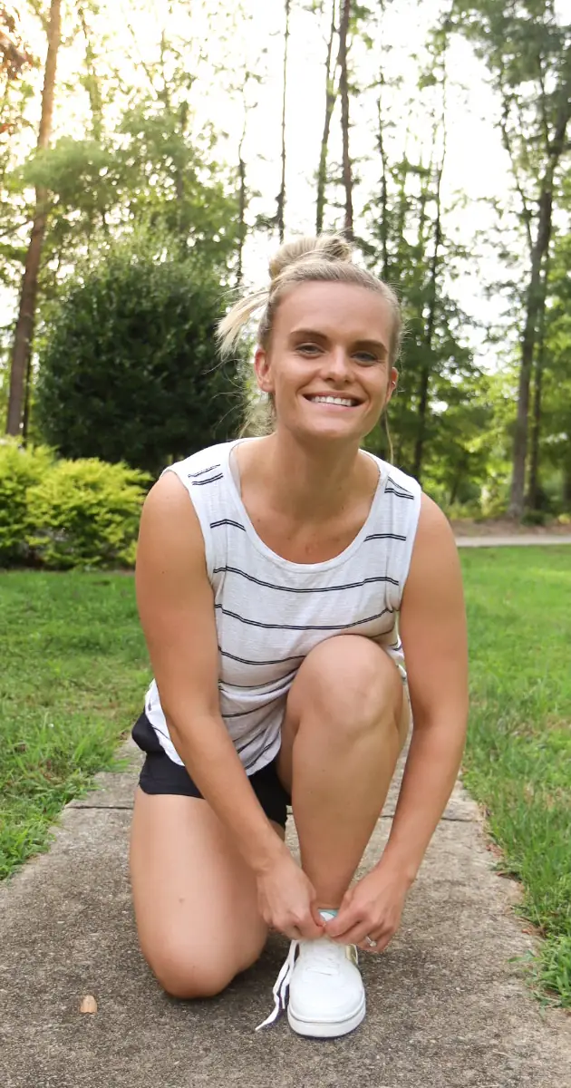 Rachel smiling at the camera while kneeling on a path to tie her running shoes.