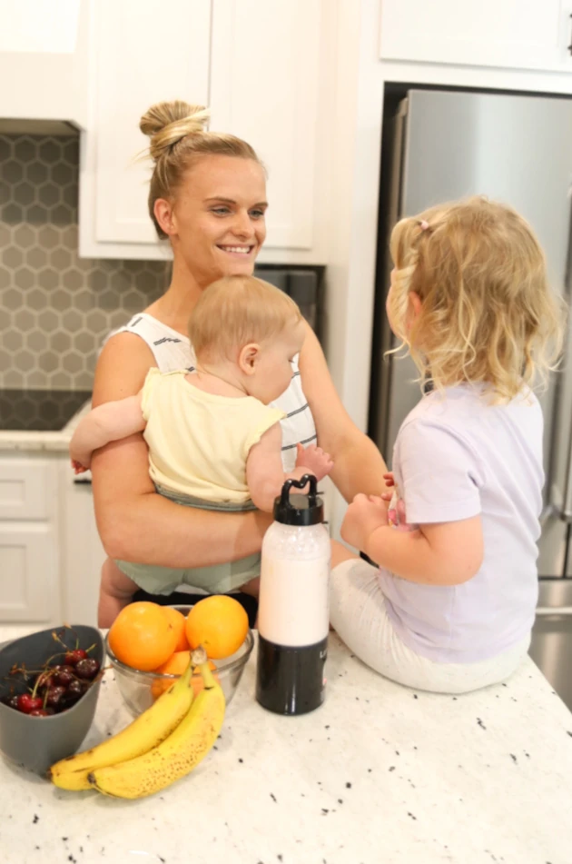 Rachel smiling and playing with her two children at the kitchen counter filled with fresh fruits.