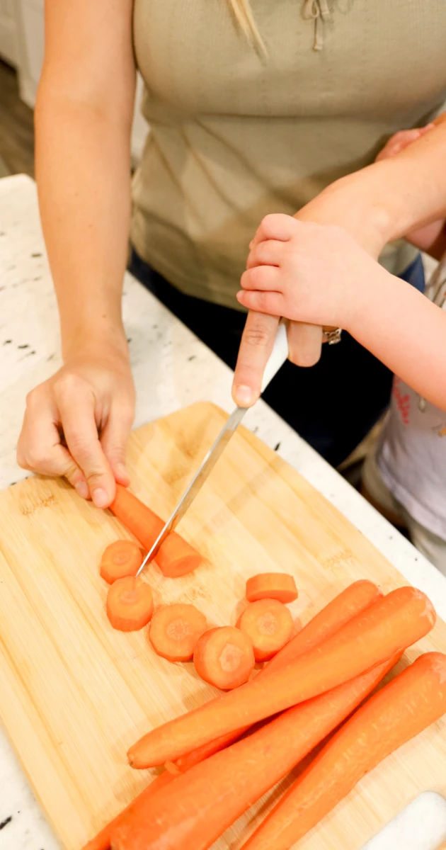 A mom cutting carrots on a cutting board while her child helps guide her hand safely.