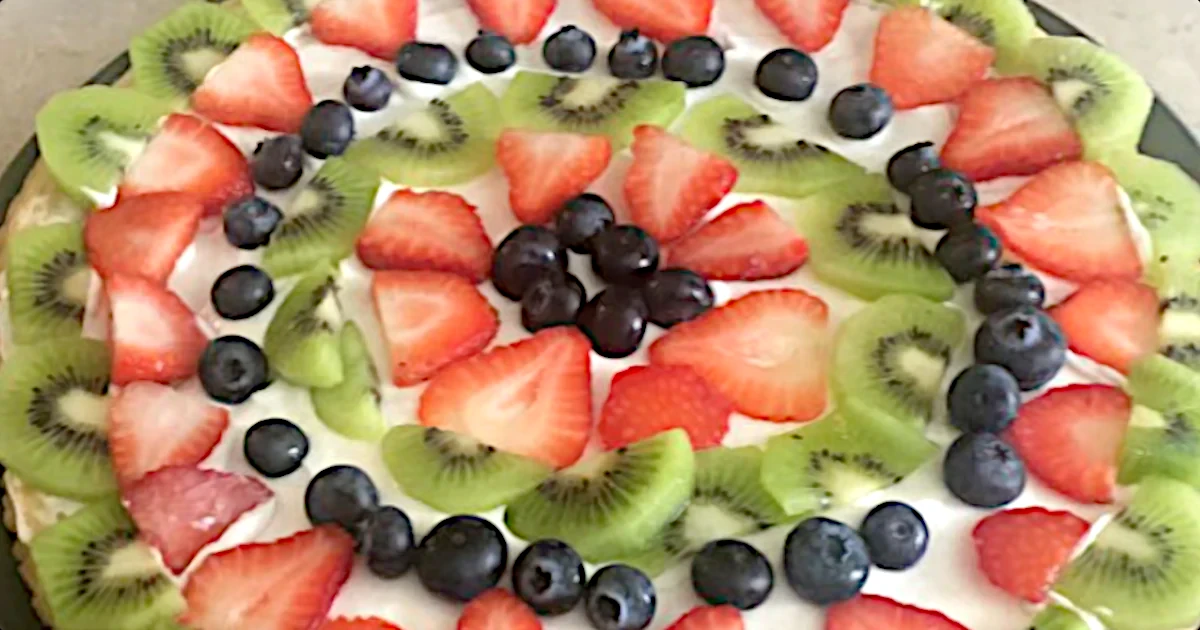 A colorful dish of fresh fruits sliced and arranged in concentric rings on a white plate, including kiwi, strawberries, and blueberries.